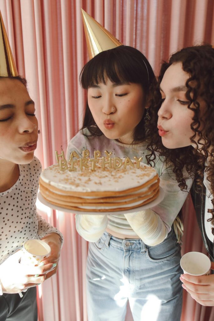 Three teenage girls blowing out candles on a birthday cake indoors.