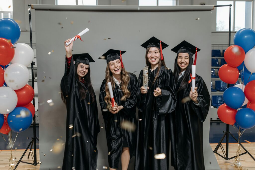 Four young female graduates celebrate indoors with caps, gowns, and diplomas.